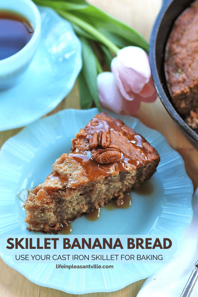 A slice of moist banana bread topped with pecans and caramel drizzle on a light blue plate, beside a cup of tea, tulips, and a cast iron skillet holding the rest of the bread.