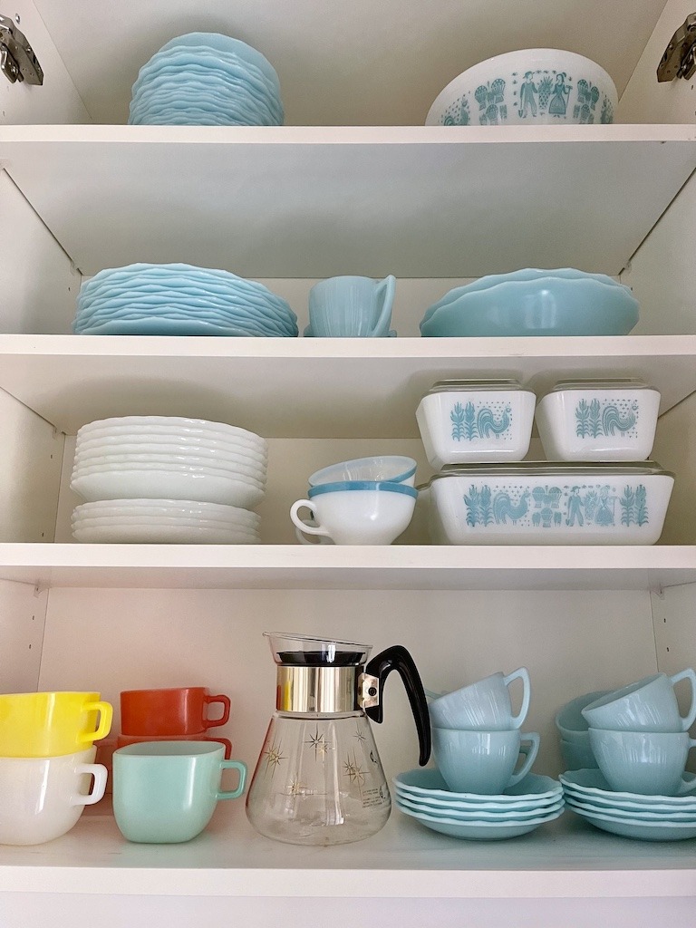 Open kitchen cabinet displaying a collection of vintage aqua and white dishes including Fire King, Pyrex Butterprint, and colourful mug sets