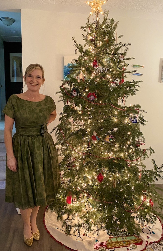 Woman smiling beside a decorated Christmas tree wearing a vintage 1960s olive green party dress with gold shoes showing that myths about thrifting prevent some beautiful purchases