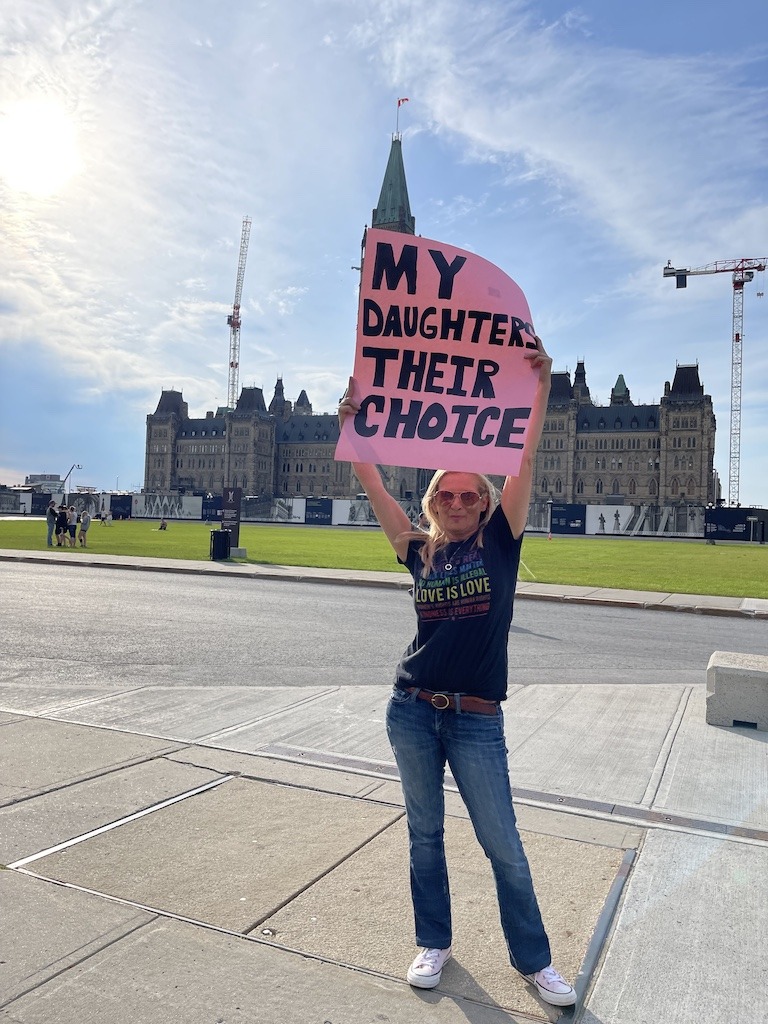 Woman holding a pink protest sign reading My Daughters Their Choice in front of Parliament Hill in Ottawa wearing thrifted flare jeans