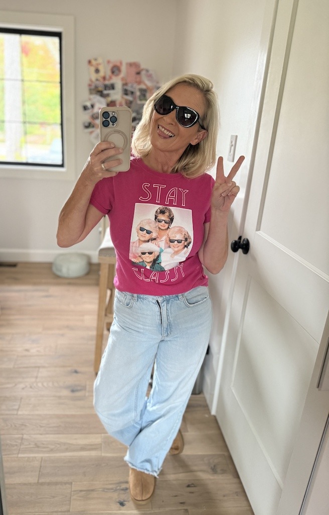 Woman taking a mirror selfie wearing a pink Golden Girls Stay Classy graphic tee with wide leg jeans and sunglasses