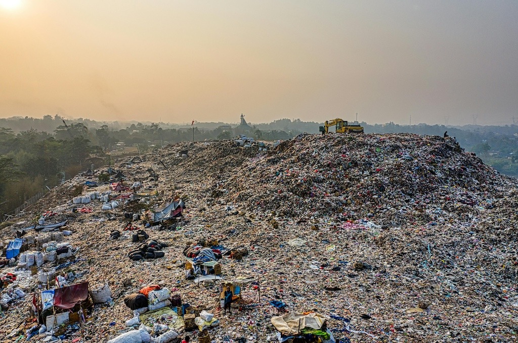 Aerial view of a massive overflowing landfill at sunset with a bulldozer on top and people sorting through waste below, illustrating the global scale of overconsumption and disposal