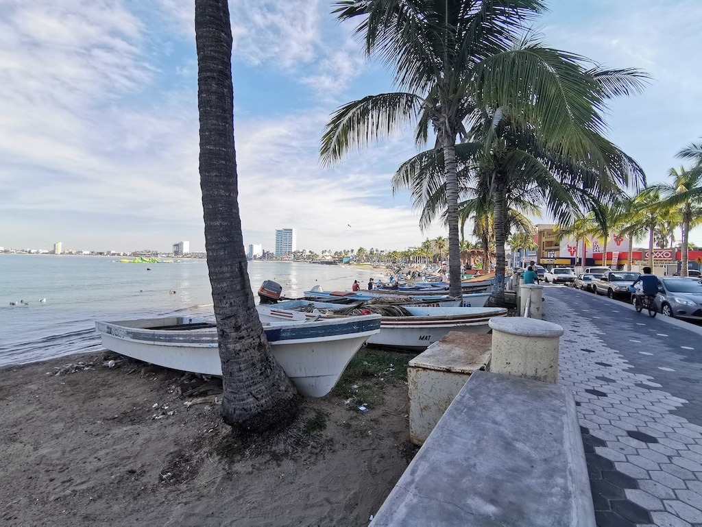 Fishing boats resting on the shore along the Mazatlán malecón with palm trees and the Golden Zone in the background.