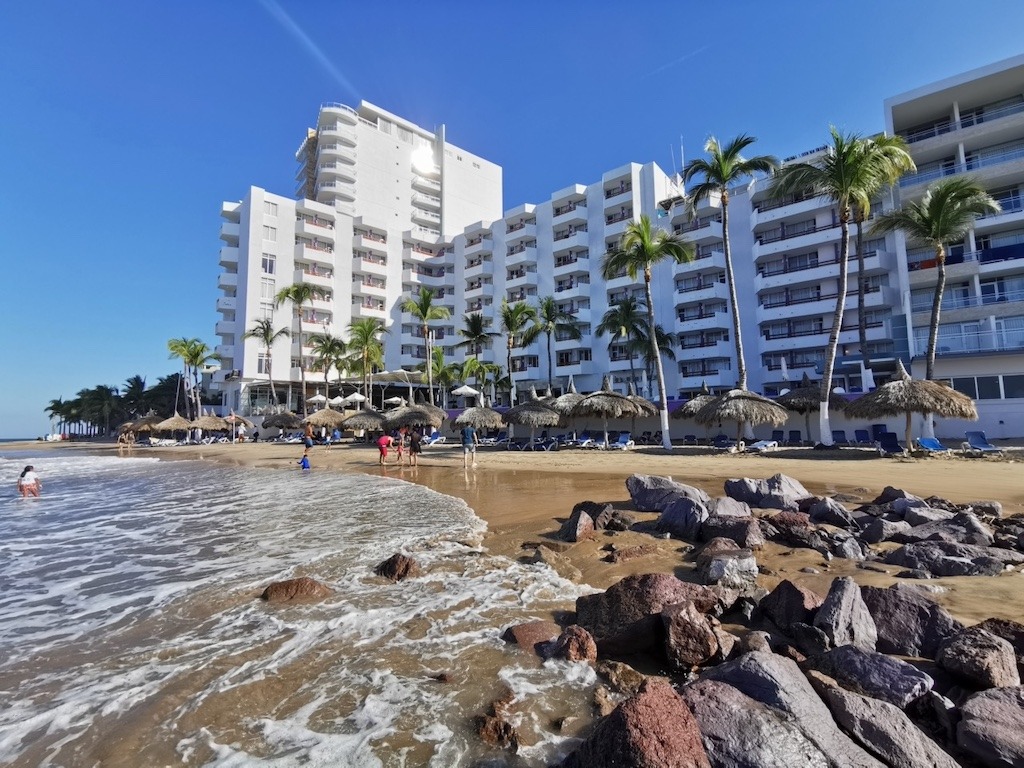 El Cid El Moro Beach Resort in Mazatlán Mexico seen from the beach with palm trees and thatched umbrellas along the Golden Zone shoreline.
