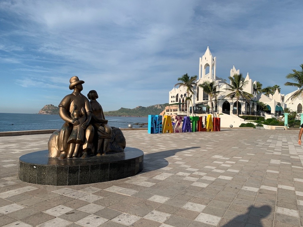 Bronze family sculpture and colourful Mazatlán sign on the malecón waterfront with a white chapel and ocean views in Mazatlán Mexico.