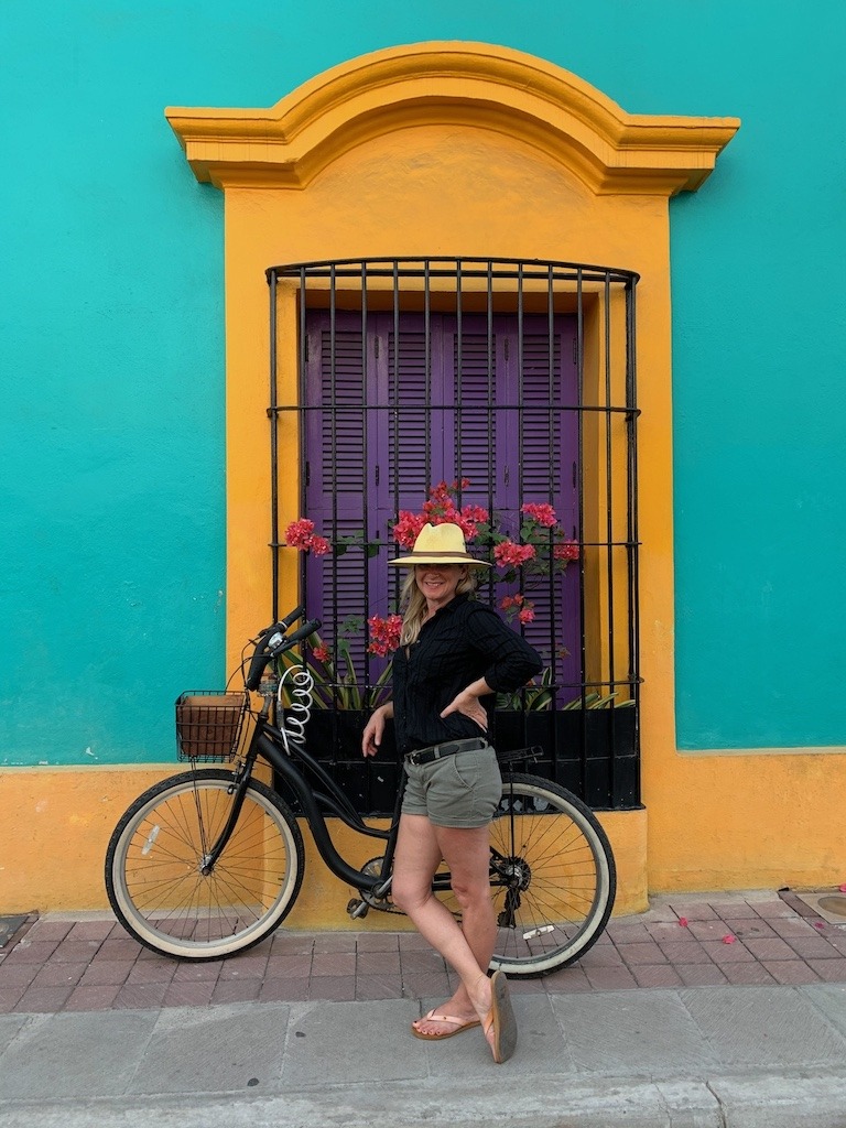 Candace standing with a bike in front of a vibrant turquoise and yellow building in Mazatlán, Mexico, wearing a straw hat and sandals.