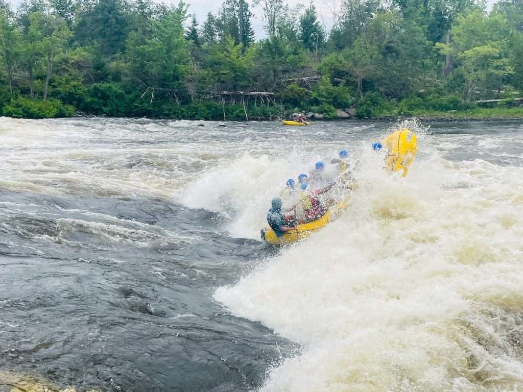 A white water rafting boat dropping into a large rapid on the Ottawa River, a kayaker following behind