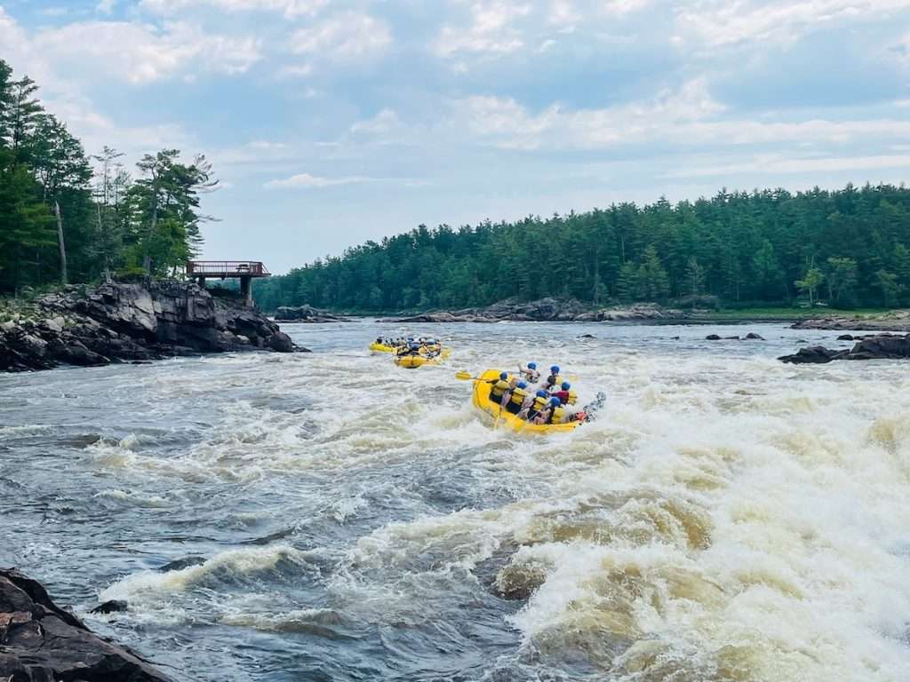 Two rafting boats running rapids on the Ottawa River with forested shoreline and a lookout platform