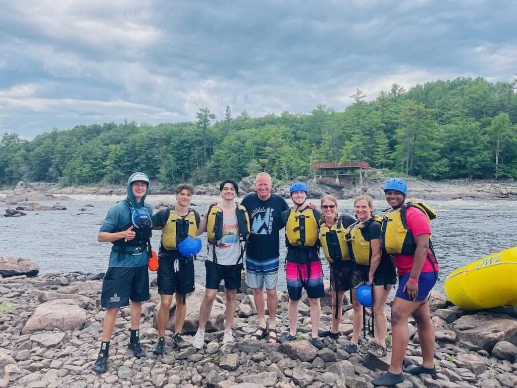 A rafting crew in life jackets posing together on the rocky shore of the Ottawa River after their run, OWL raft beside them