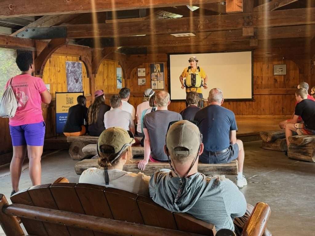Guests seated on log benches watching a guide demonstrate a life jacket during the pre-trip safety briefing at OWL Rafting