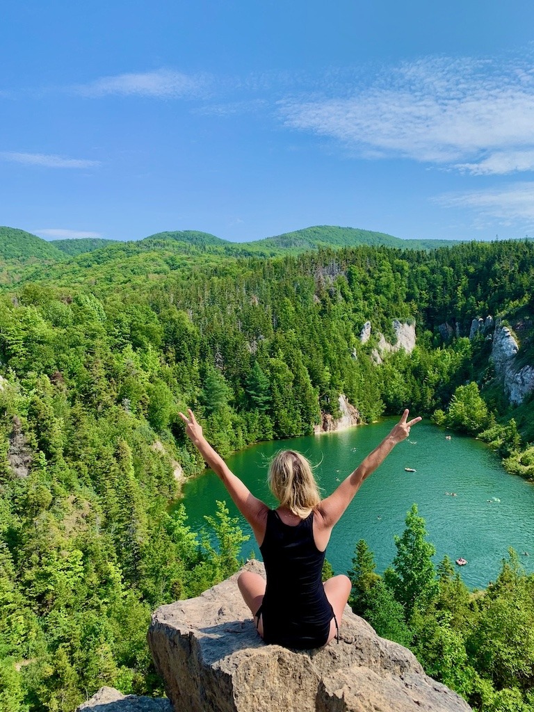 Woman sitting on a cliff overlooking a lake and forest in Cape Breton, Nova Scotia, celebrating nature and adventure.