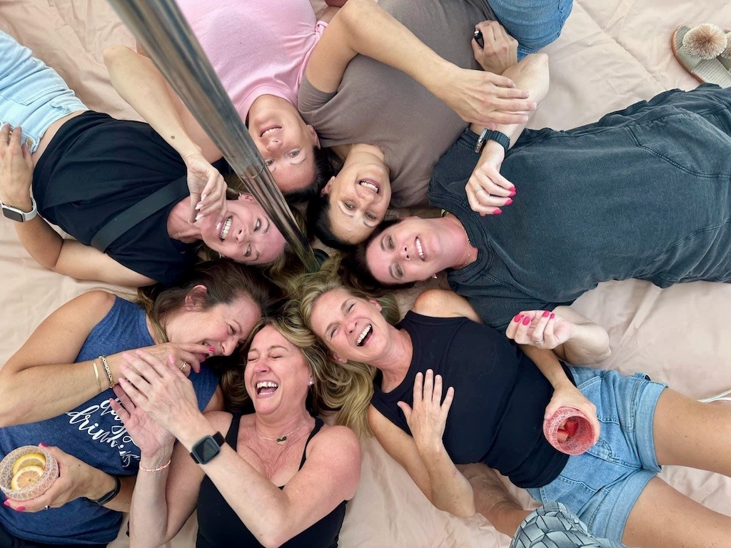 A group of women lying in a circle on a blanket inside a glamping tent, laughing and holding drinks, capturing the joy of a carefree girls’ getaway in nature.