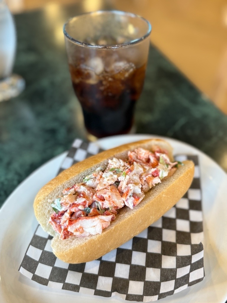 Classic East Coast lobster roll served on a checkered napkin with a glass of cola, perfect festival fare in Cape Breton.