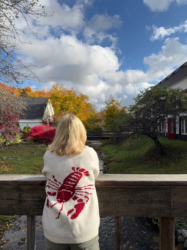 Woman in a white sweater with a red lobster design looking over a creek at Glenora Inn and Distillery dressed in peak fall colours. Plan your Cabot Trail Fall Road Trip.
