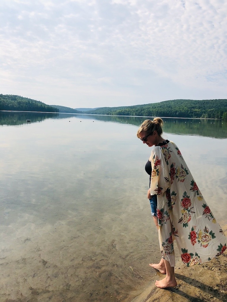 Woman standing barefoot at the edge of a still lake in cottage country, present and unhurried in the natural world