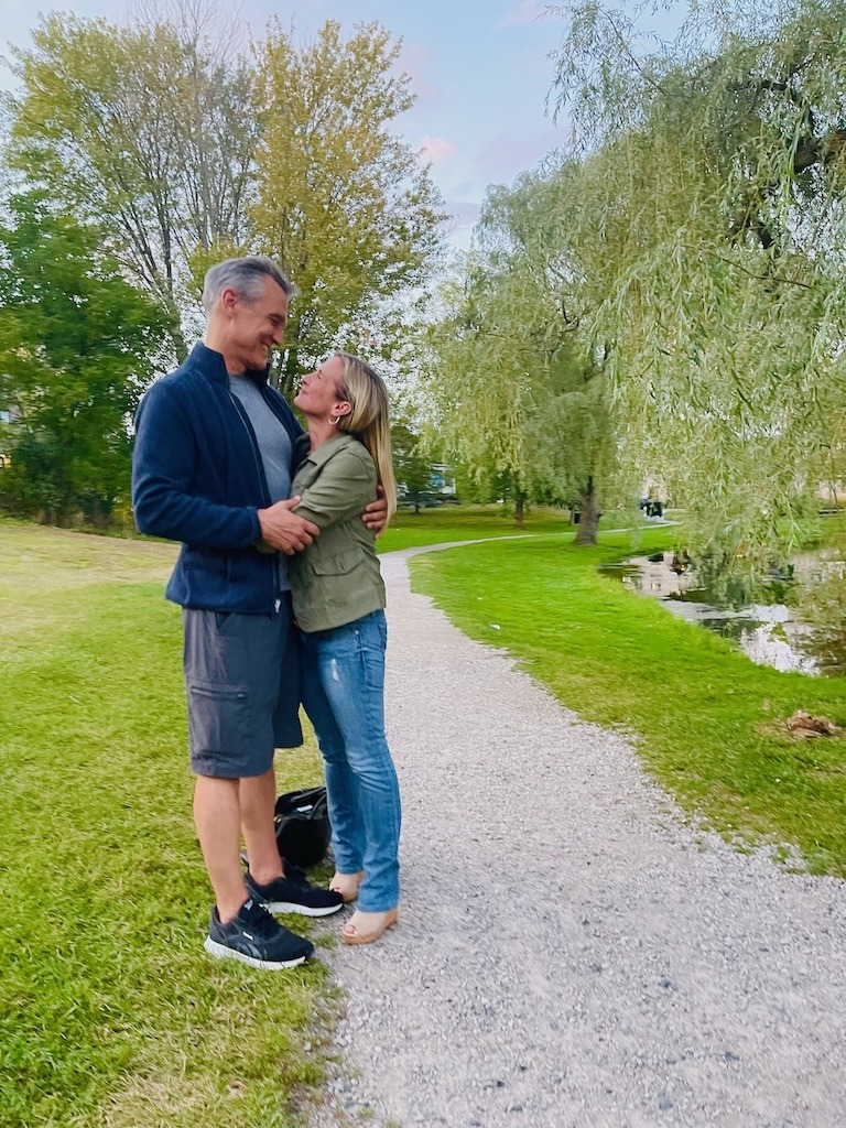 A couple laughing together on a park path surrounded by willow trees, the kind of real-world human connection that no dating app can replicate