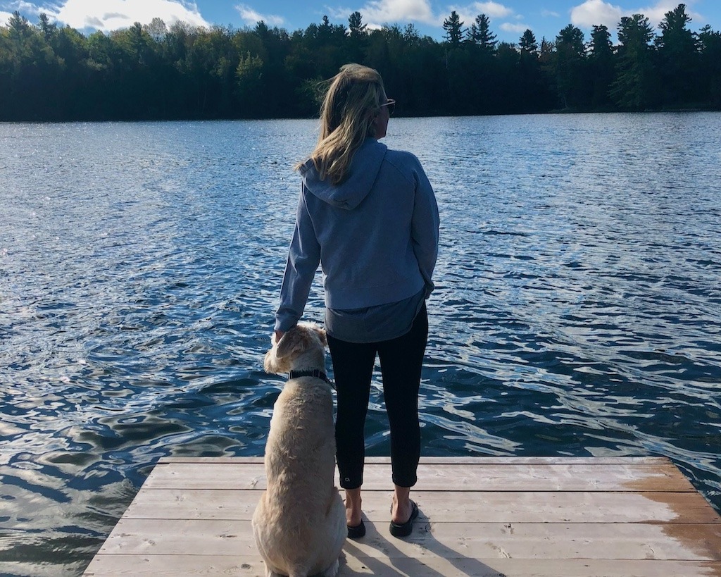 Woman and her dog standing on a wooden dock looking out at a calm Canadian lake and forest, a quiet act of resistance against extinction of experience.