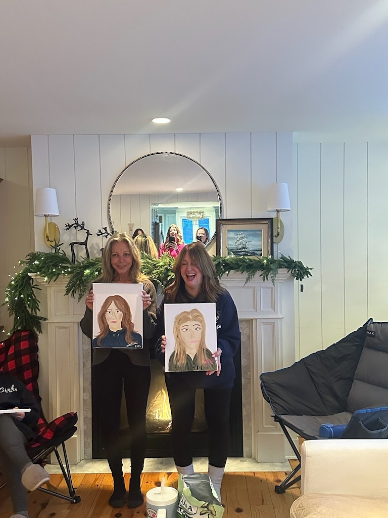 Two women holding up their painted portraits during a Crystal Beach Ontario winter girls weekend reveal