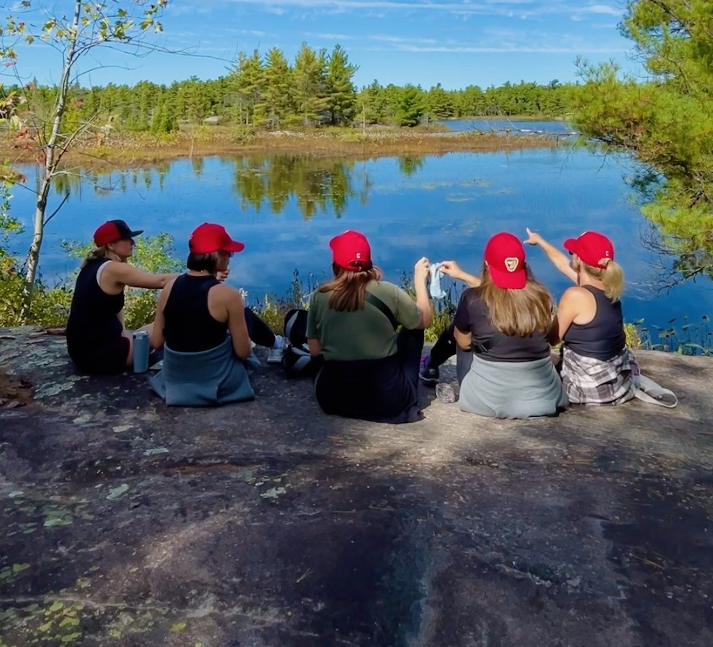 Five women sitting together on a rock overlooking a Canadian lake, connecting in real life through Girl Trips