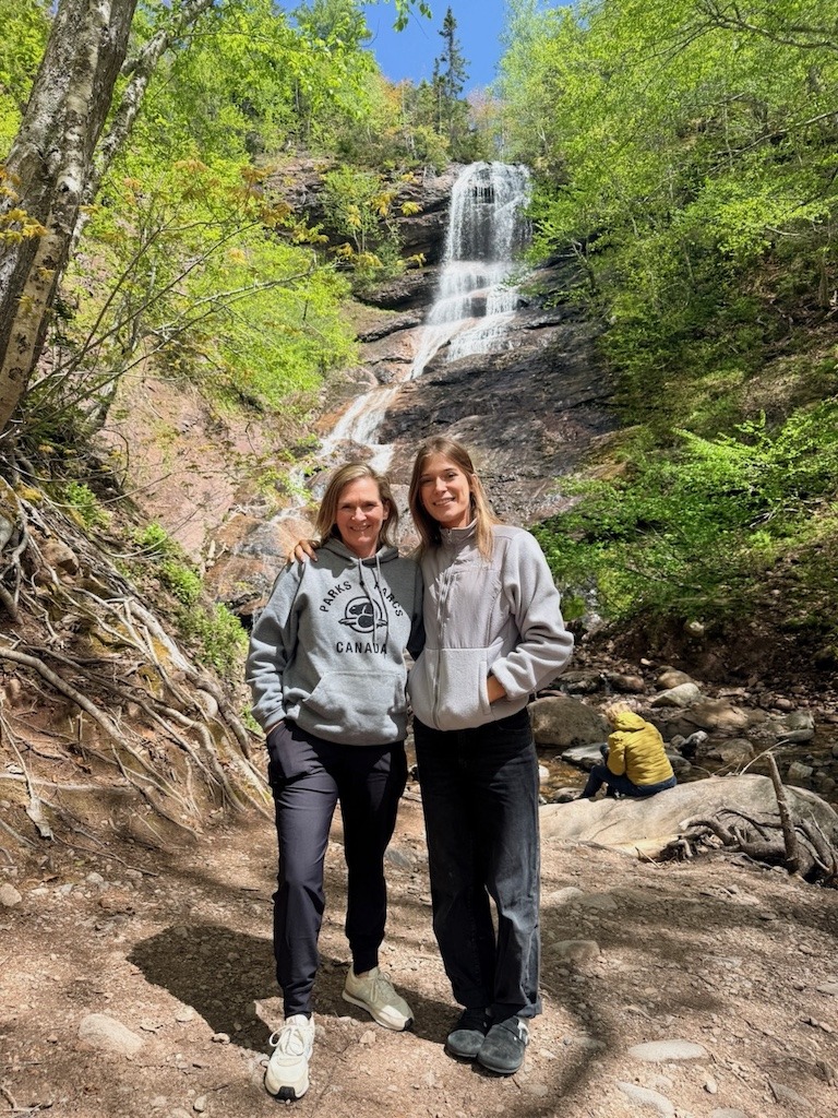 Two women in Parks Canada hoodies chasing waterfalls in Cape Breton in front of a tall spring waterfall