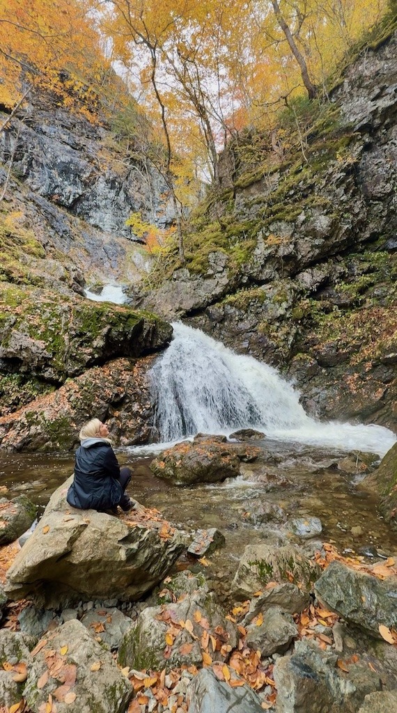Woman sitting on a rock below a waterfall surrounded by fall colour in Cape Breton