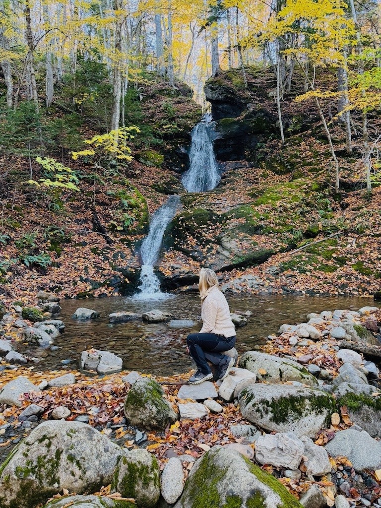 Woman kneeling beside a tall two tiered waterfall framed in autumn leaves in Cape Breton
